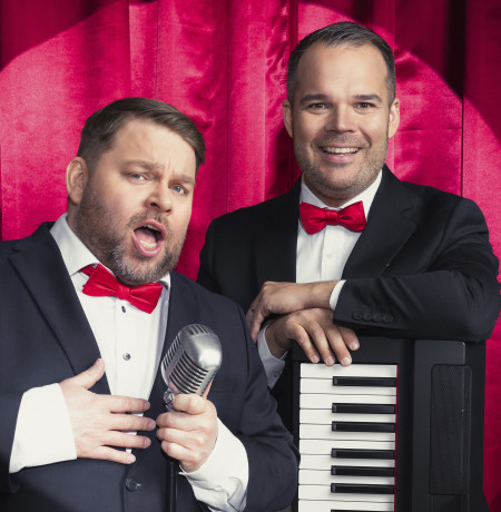 Näyttelijät Mikko ”Tökä” Töyssy ja Mikael ”Micke” Konttinen puvut päällä punaiset rusetit kaulassa punaisen esiripun edessä. 