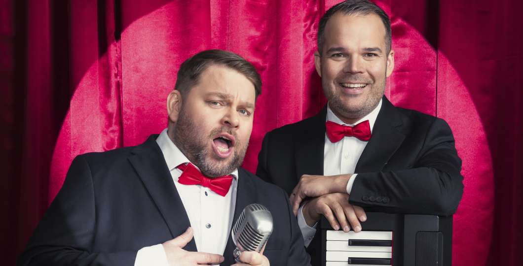 Näyttelijät Mikko ”Tökä” Töyssy ja Mikael ”Micke” Konttinen puvut päällä punaiset rusetit kaulassa punaisen esiripun edessä. 